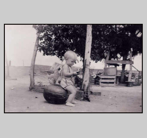 <adrienne sitting on a watermellon. The door to grandma's cellar can be seen in the background>